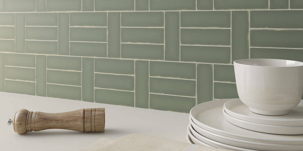 Sage green kitchen backsplash tile in a basketweave pattern above a light countertop, styled with a wooden pepper mill and stacked white plates and bowl.