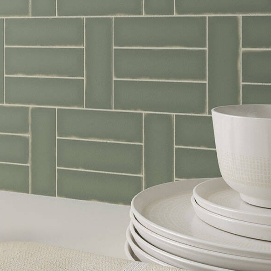 Sage green kitchen backsplash tile in a basketweave pattern above a light countertop, styled with a stacked white plates and bowl.