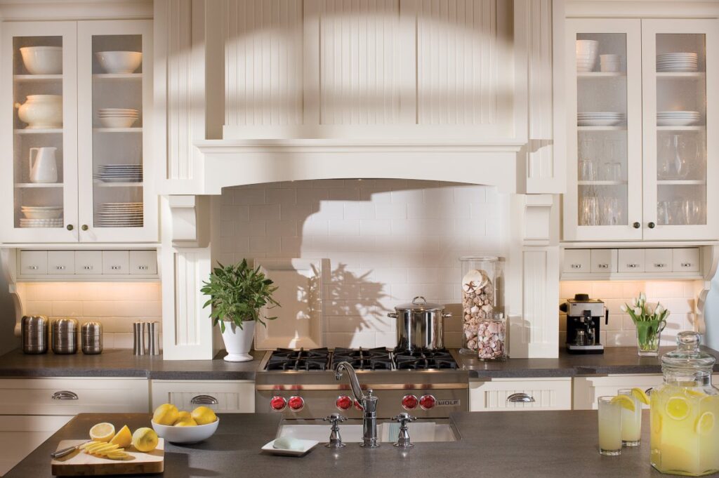 A beautifully crafted space featuring Dura Supreme antique white kitchen cabinets offered at JC Huffman, highlighting a custom beadboard range hood, classic glass display doors, and elegant dark countertops.