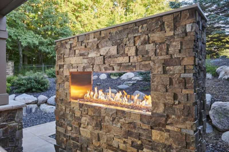 Outdoor gas fireplace with a long, horizontal flame set into a natural stone wall beside a landscaped patio.